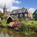 A village with green and black wooden houses and a churchtower, behind a canal and flowers in a garden