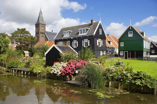 A village with green and black wooden houses and a churchtower, behind a canal and flowers in a garden
