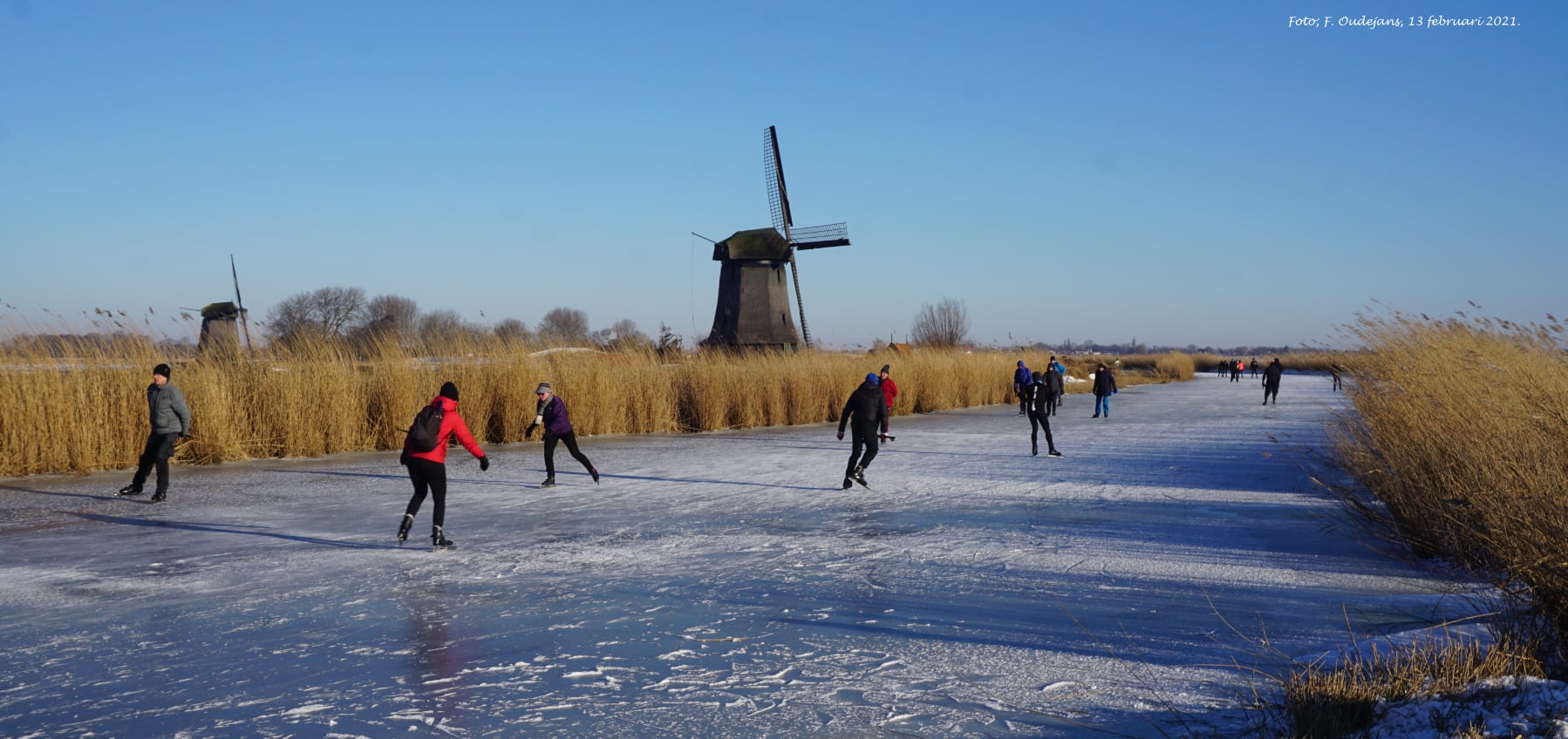 people ice skating on a canal near a traditional windmill under a clear blue sky
