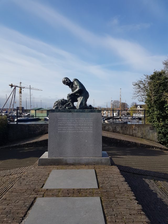 a statue of a boy with his finger in the dike with boats at the background