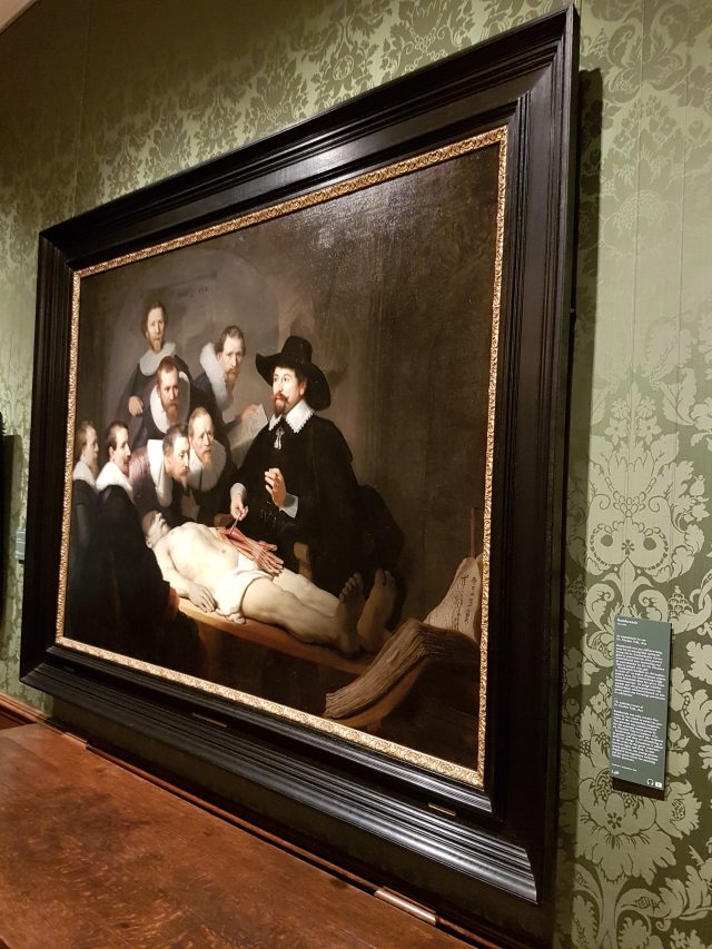 A painting on a wall with a group of people in black clothes around a white body lying on a table