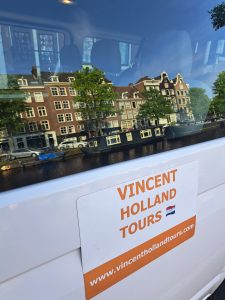 a door of a white van with a sign with orange text and canal houses can be seen through in window as reflection